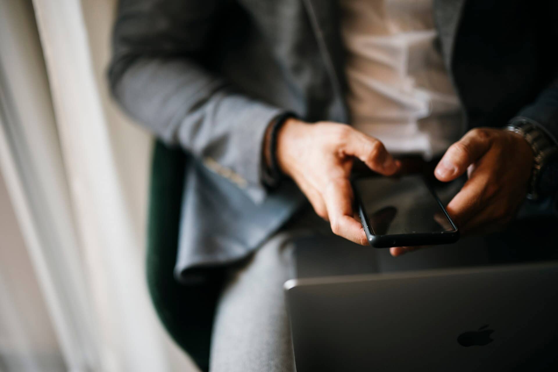 Homem sentado segura o celular com as duas mãos ao lado de um notebook
