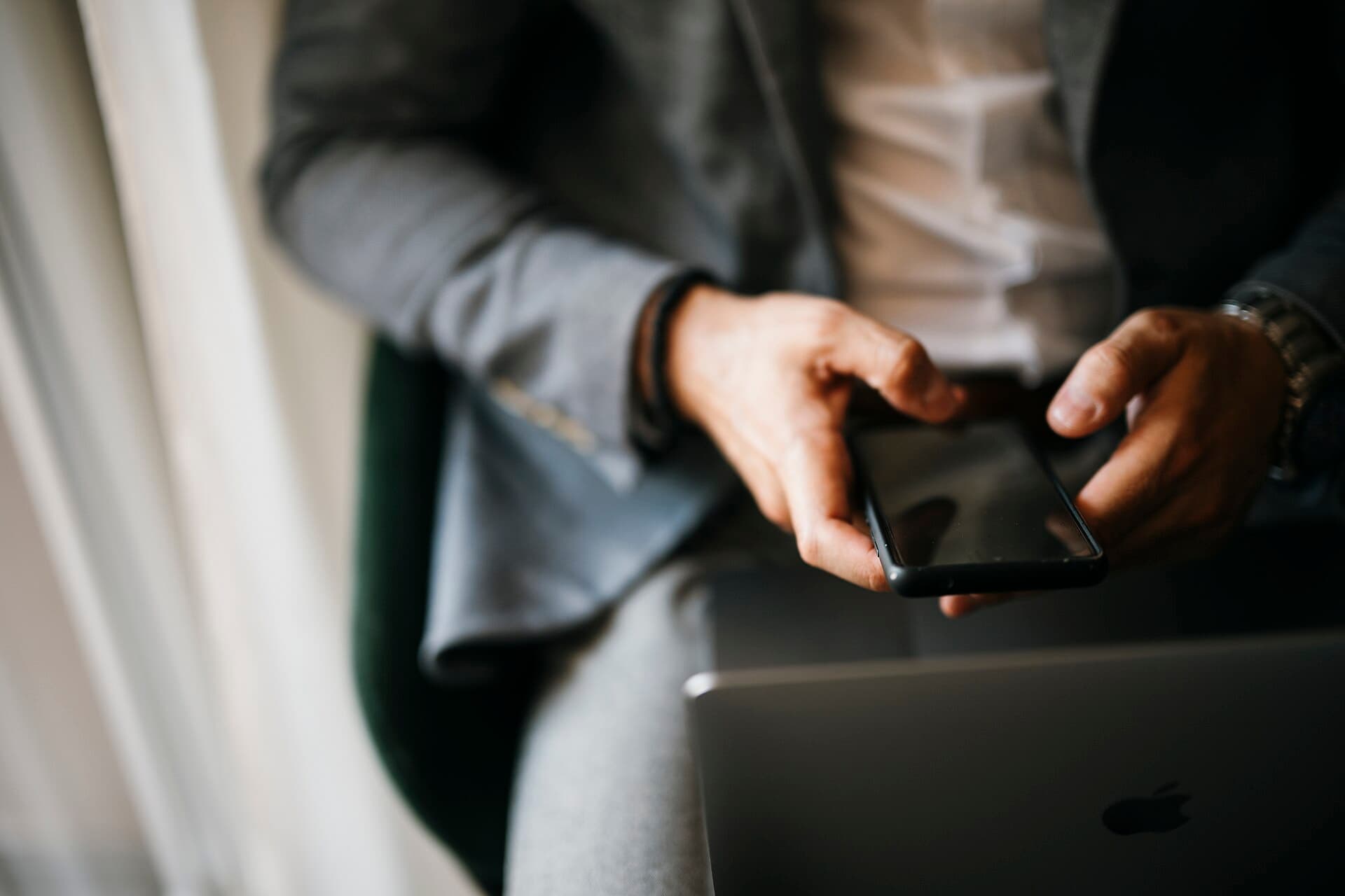 Homem sentado segura o celular com as duas mãos ao lado de um notebook
